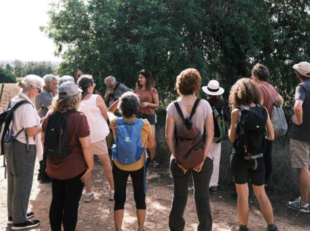 Un grup de persones amb motxilla, en rotllana en un camí de terra, escoltant na Malena sota uns arbres